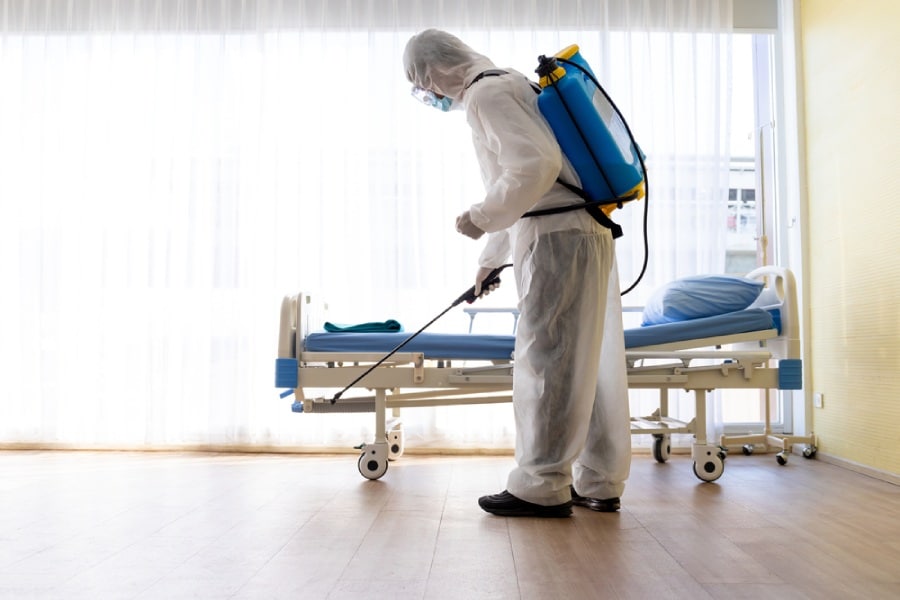 Hospital worker in PPE cleans and disinfects hospital bed per OSHA exposure control plan.