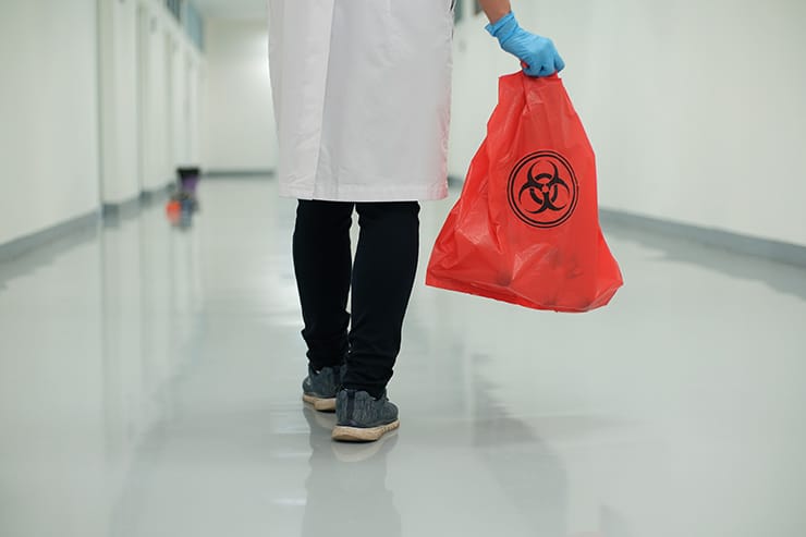 Doctor in white coat carries a red bag of medical waste with biohazard symbol to the trash bin.