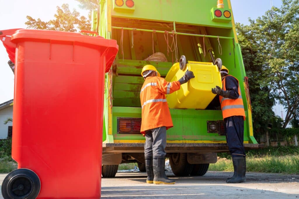 Two garbage men filling up a dump truck with office waste from a red garbage bin.